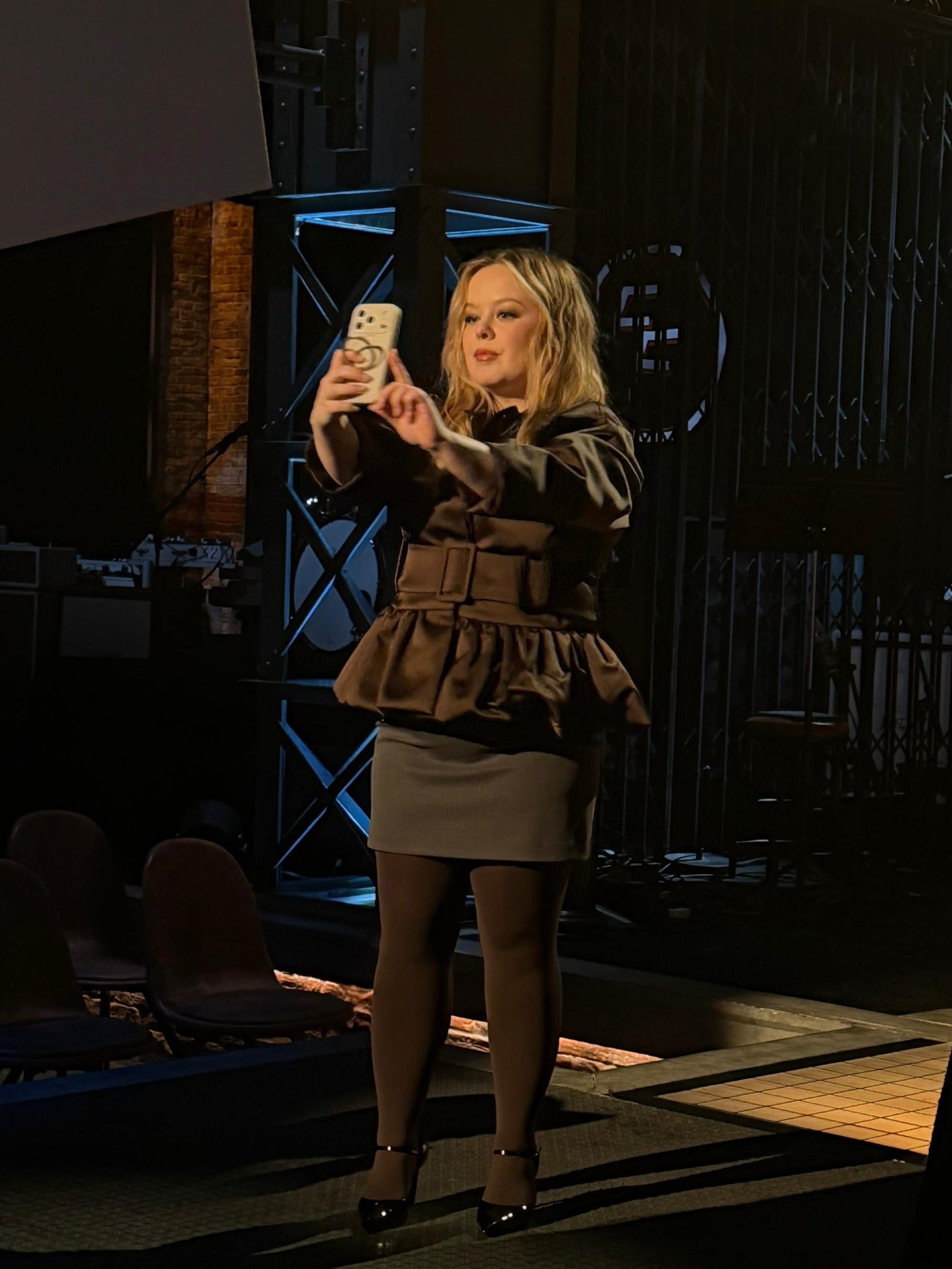 Nicola Coughlan on a stage taking a photo with a smartphone, surrounded by people and a large screen.