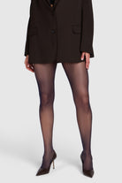 A model from the waist down wearing sheer black tights, a dark brown blazer, and dark brown pointed-toe heels against a light gray studio background.