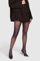 A model wearing sheer black tights, a dark brown blazer, and dark brown pointed-toe heels against a light gray studio background.